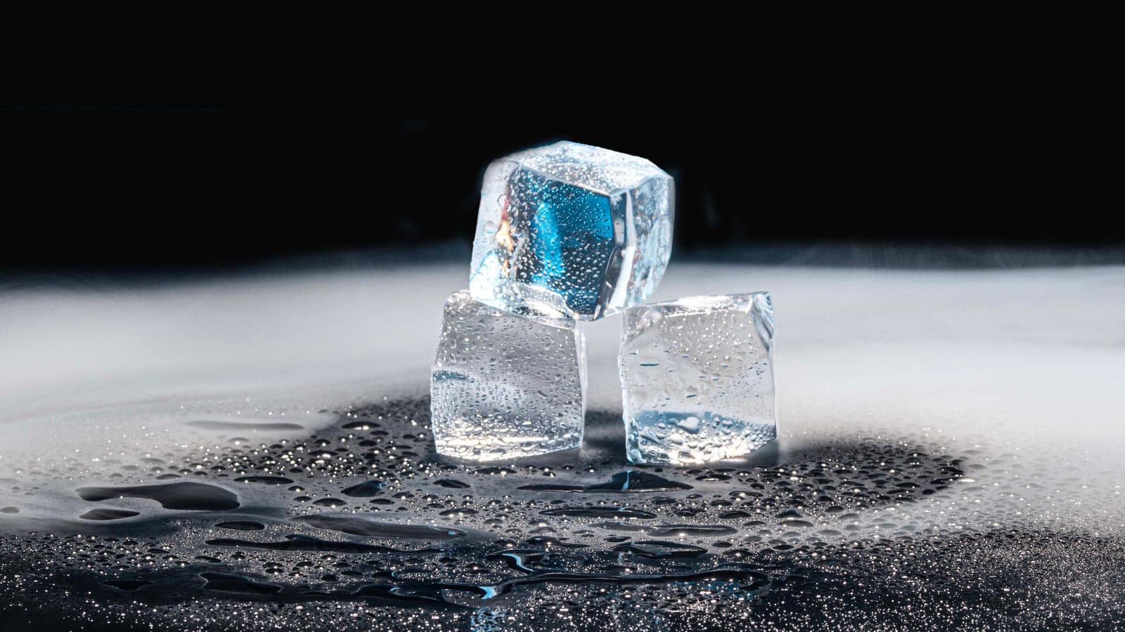 Three ice cubes melting on a black surface with water droplets
